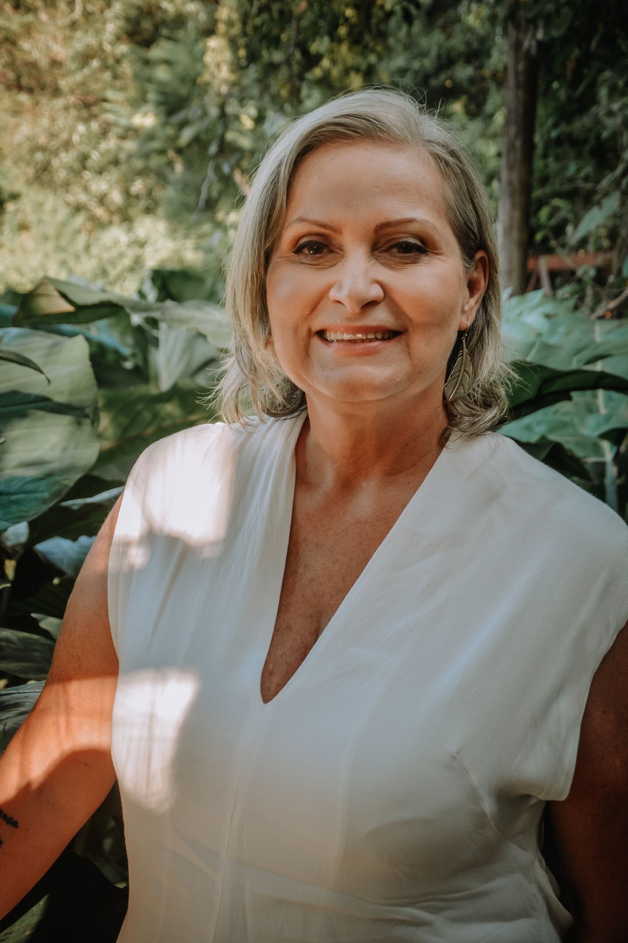 Smiling person in a white top standing in front of lush green foliage.