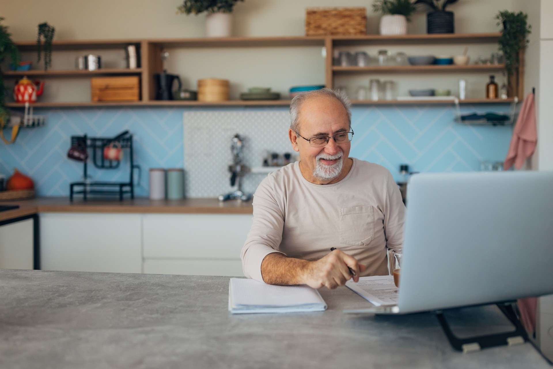 Senior adult man with eyeglasses uses his laptop for further education through e-learning. He sits comfortably at the kitchen table, taking advantage of technology to advance his knowledge